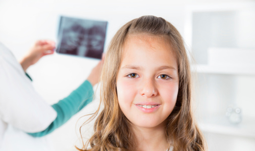 Child smiling in a dental office while a clinician reviews an image in the background